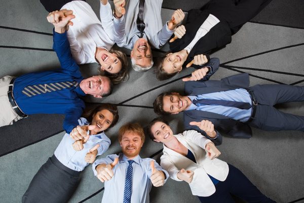 Eight smiling at camera business people lying on floor in circle and showing thumbs up. Top view.